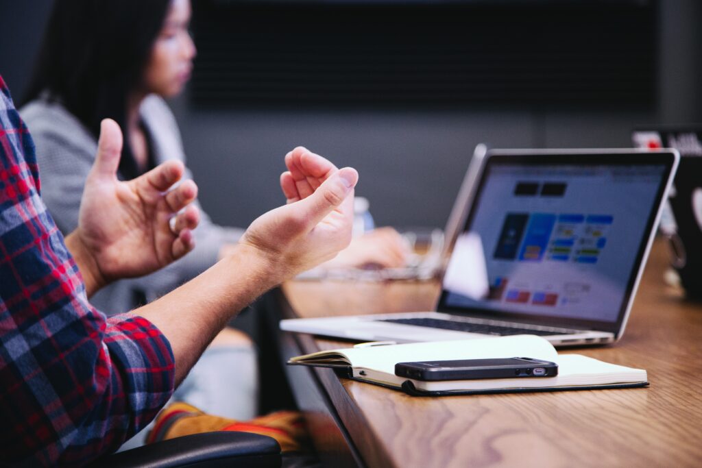 Two people sitting at a table in front of a laptop. One person is a white man, who is gesturing at the computer. The other is an asian woman looking at the computer.