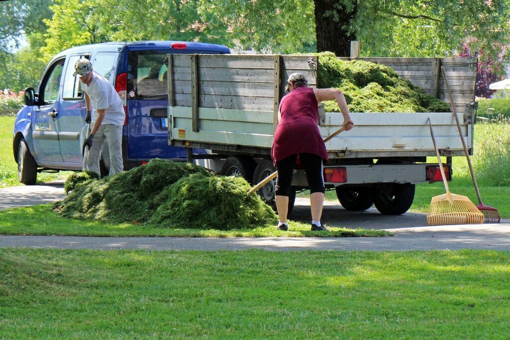 Two white people shoveling loose yard debris next to a truck.