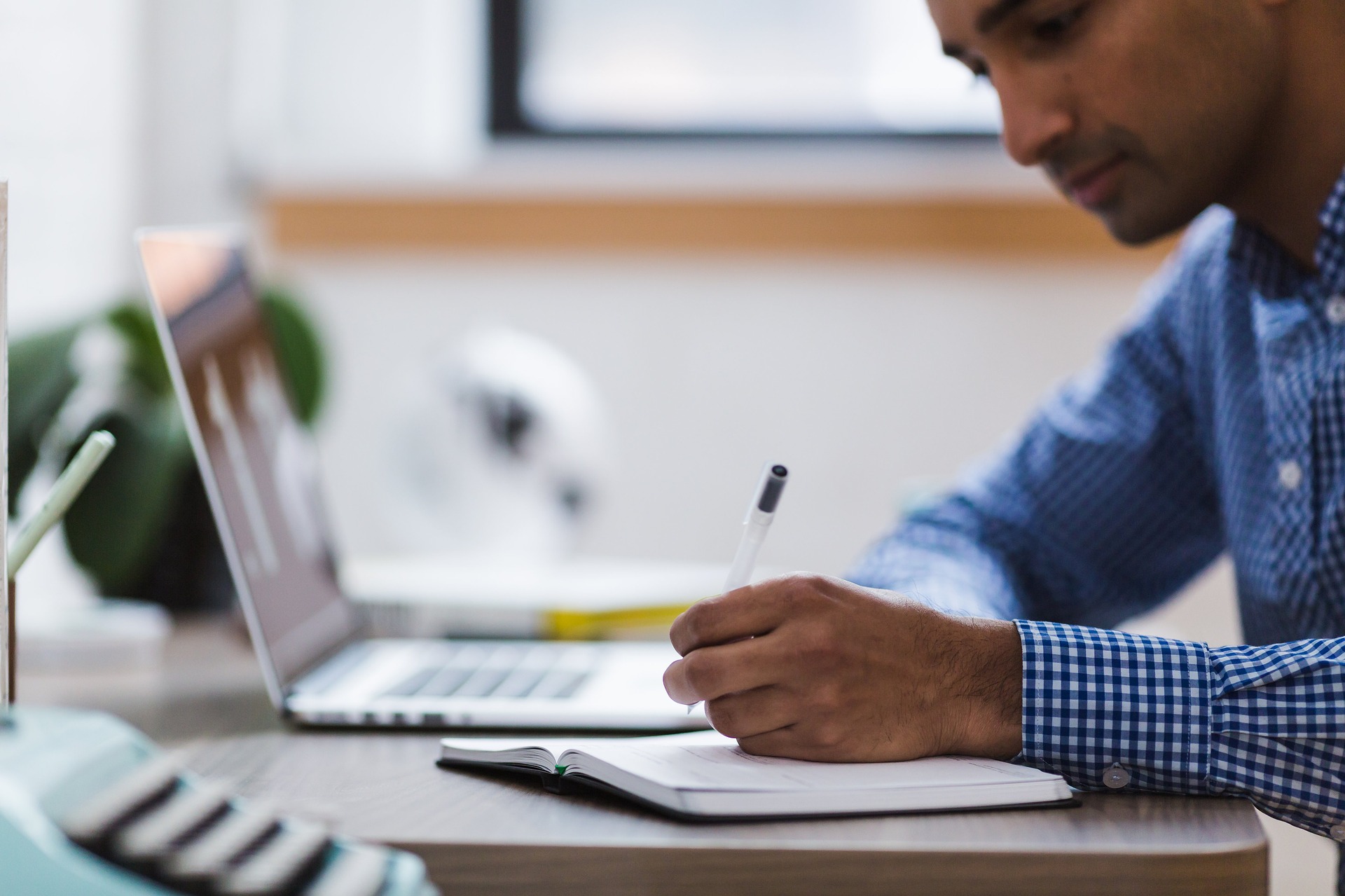 A man with a brown skin tone sits in front of a laptop. He is writing in a notebook.