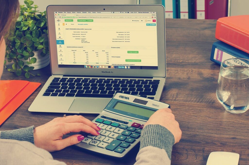 A white person sitting at a computer with a calculator. The computer is showing a spreadsheet of tax values.