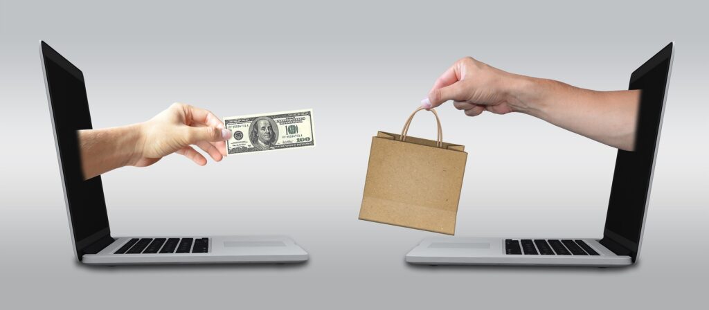 Two hand sticking out from two laptop screens facing each other. One hand is holding a dollar bill, the other is holding a shopping bag.